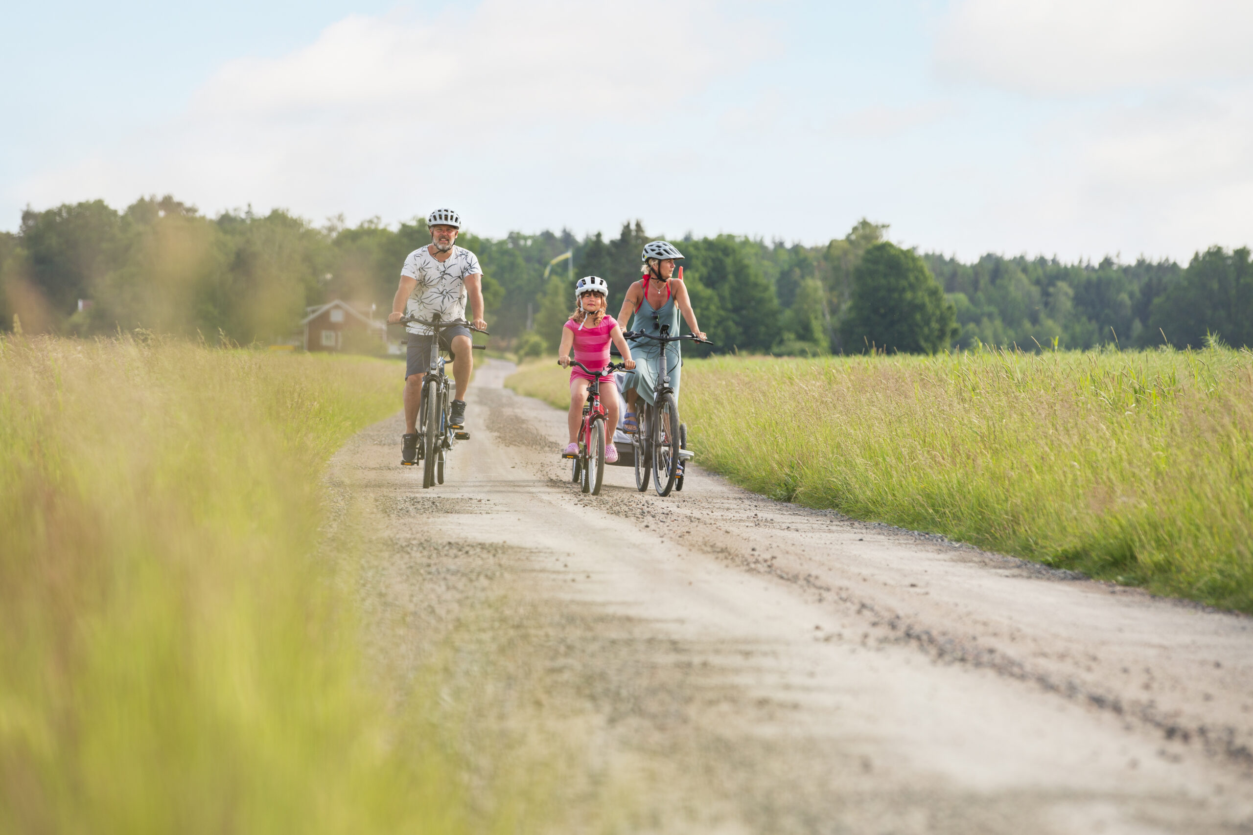 En familj med pappa, mamma och liten flicka cyklar på en grusväg omgiven av öppna fält.