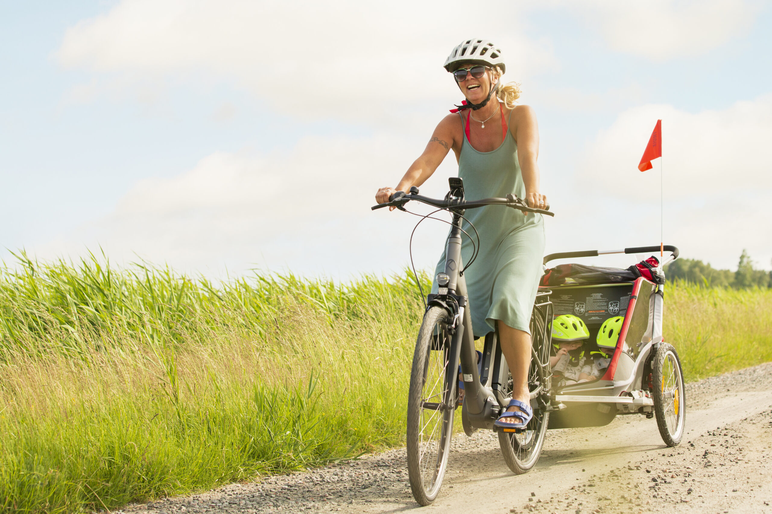 Kvinna cyklar på grusväg med cykelkärra med två barn i.